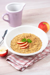 Bowl of oatmeal with sliced apples on a checkered cloth, next to a purple pitcher and apple slices.